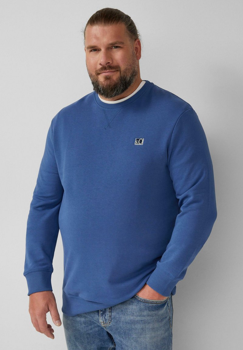 Felpa blu con collo a girocollo a costine e maniche cuffiate, caratterizzata da un piccolo logo ricamato sul petto, abbinata a jeans azzurri chiari.