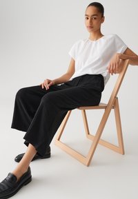 Witte top met korte mouwen, zwarte wijde broek en zwarte instappers; zittend op een houten opvouwstoel. Glad textuur, casual fit.