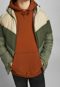 Pikowana kurtka w zielono-beżowe bloki kolorystyczne, z kapturem i zapięciem na zamek, noszona na rdzawej bluzie. Teksturowany materiał.
