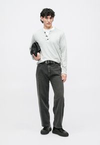 Hombre con una camiseta henley gris claro y jeans negros sosteniendo una chaqueta de cuero negra frente a un fondo blanco liso.