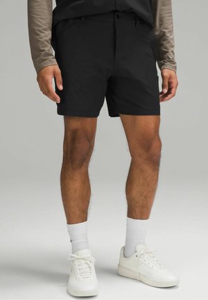 Mężczyzna ubrany w czarne spodenki, białe trampki i białe skarpetki, stojący z ramionami rozluźnionymi na tle jednolitego jasnoszarego tła.