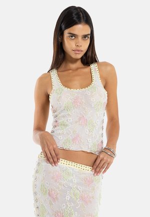 Vrouw draagt een mouwloze pastelbloemen kanten top met met studs afgewerkte rand en bijpassende rok, poseert met handen bij de taille tegen een witte achtergrond.