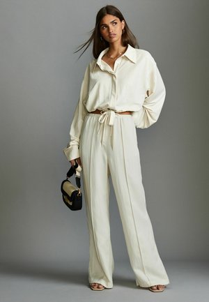 Krémová oversized košile s límcem, spárovaná s odpovídajícími kalhotami s vysokým pasem. Modelka drží černou a krémovou kabelku.