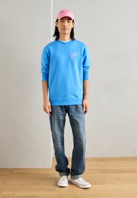 Lichtblauwe sweatshirt met logo, gecombineerd met klassieke blauwe spijkerbroek en witte sneakers. Het model draagt een roze pet en staat tegen een neutrale achtergrond.