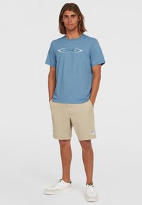 Blå meleret t-shirt med et hvidt logo, parret med beige shorts med en logo-patch. Hvide sneakers fuldender outfittet.