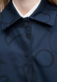 Κοντινό πλάνο μπλε σκούρου πουκαμίσου με μαύρα αφηρημένα κυκλικά σχέδια, κουμπωμένο μπροστά, και λευκό V-λαιμοκάλυμμα που φαίνεται στο γιακά.