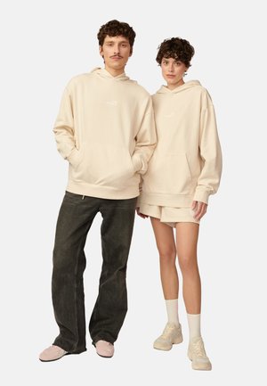 Beżowe bluzy z kapturem, z kieszeniami z przodu, w oversize'owym kroju, z ściągaczami przy mankietach i u dołu. Model w ciemnych dżinsach, drugi w mini spódnicy i skarpetkach.