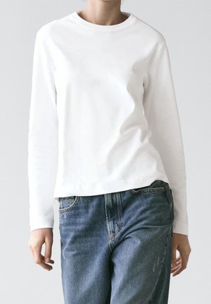 Personne portant une chemise blanche à manches longues simple, légèrement rentrée dans un jean bleu taille haute, sur un fond clair.