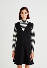 Vestido negro sin mangas con escote en V, llevado sobre un cuello de tortuga a rayas en blanco y negro. Tejido suave, corte relajado y diseño hasta la rodilla.