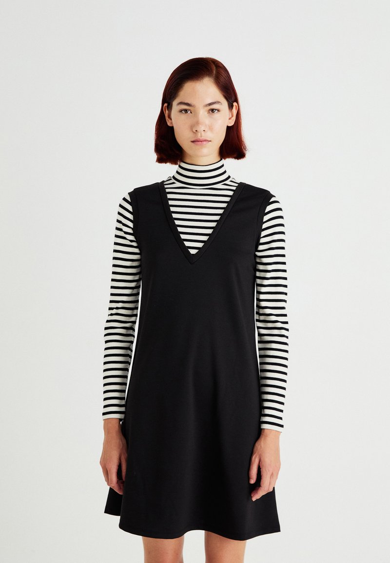 Vestido negro sin mangas con escote en V, llevado sobre un cuello de tortuga a rayas en blanco y negro. Tejido suave, corte relajado y diseño hasta la rodilla.