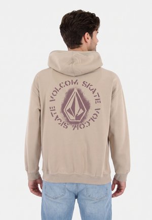 Hombre con sudadera con capucha beige con el logo "Volcom Skate" impreso en púrpura en la espalda, combinado con vaqueros azul claro, de espaldas.
