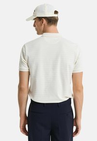 Polo crème avec des rayures horizontales subtiles, manches courtes et coupe décontractée, associé à un pantalon bleu marine foncé et une casquette claire.