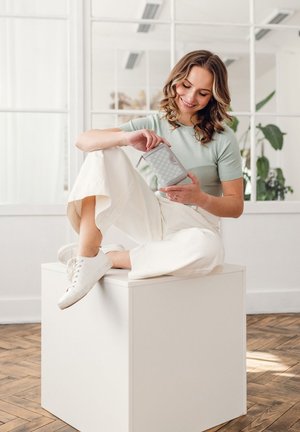 Light gray wallet with a hexagonal pattern held by a person in a mint green top and white wide-leg trousers, sitting on a white cube.