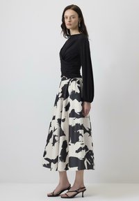 Woman in black long-sleeve ruched top, black-and-white abstract print midi skirt, black heeled sandals, and pearl drop earrings, standing sideways.