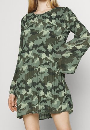 Kvinde iført en langærmet, plisseret kjole med grøn og mørkegrøn camouflageprint og en løs, flydende pasform.