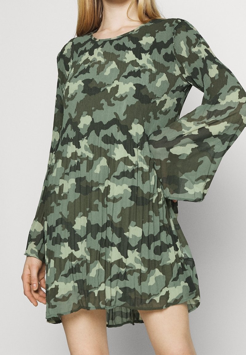 Femme portant une robe à manches longues, plissée, imprimée camouflage vert et vert foncé, avec une coupe ample et fluide.