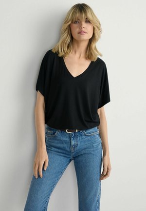 Blondynka o włosach do ramion, ubrana w czarną bluzkę z dekoltem w serek i niebieskie dżinsy, stoi na tle jasnej, jednolitej ściany.