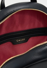 Interno di uno zaino in pelle nera con fodera in tessuto rosso scuro e tasca con cerniera. Dettagli in metallo dorato e etichetta del marchio visibili.