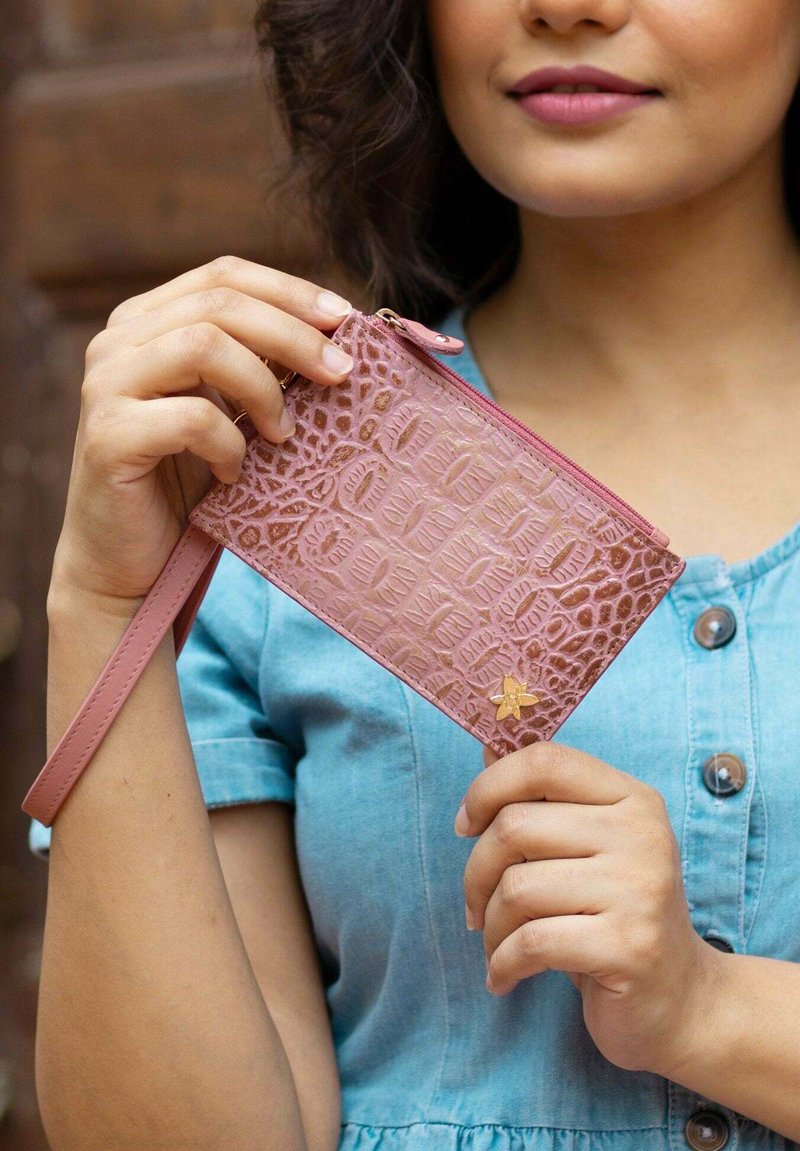 Lichtroze portemonnee met krokodillenstructuur, ritssluiting en polsband. Bevat een kleine gouden bloemenaccent. Gladde leren materiaal.
