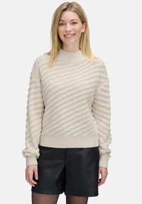 Lächelnde Frau mit glattem blonden Haar, die einen strukturierten cremefarbenen Strickpullover und schwarze Lederhosen trägt, vor einem schlichten Hintergrund.