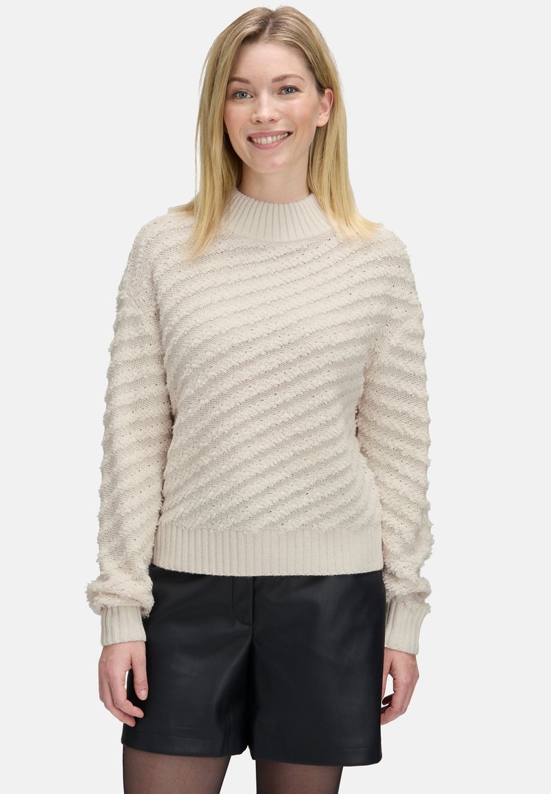 Lächelnde Frau mit glattem blonden Haar, die einen strukturierten cremefarbenen Strickpullover und schwarze Lederhosen trägt, vor einem schlichten Hintergrund.