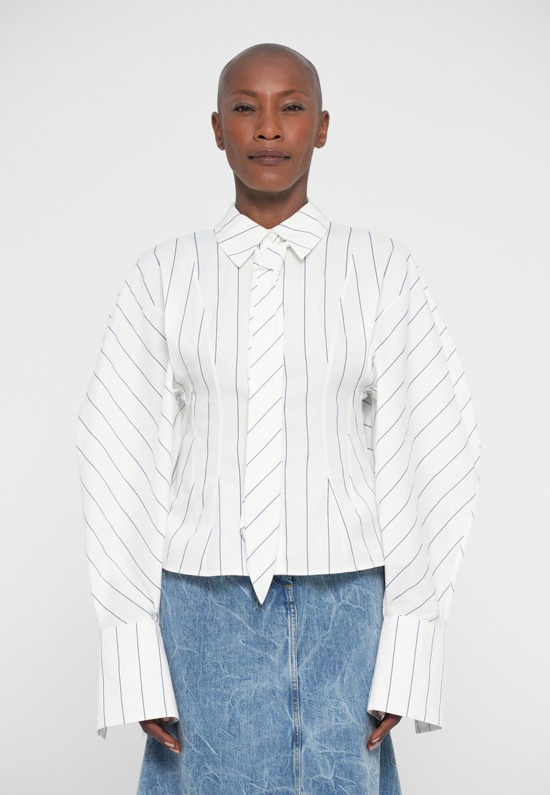 Woman with shaved head wearing a white blouse with thin vertical stripes and an attached necktie, paired with a blue denim skirt.