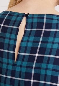 Zadní část krku s modro-zeleným kostkovaným oděvem, který má malý černý knoflík zapínající otvor v límci.