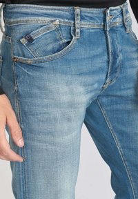 Jean en denim bleu clair avec une texture délavée, doté d'un design à cinq poches, de coutures contrastantes et d'une petite étiquette de marque sur la taille.
