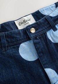 Sötét farmeranyag nagy világoskék pöttyökkel. Sárgaréz gombos záródás a "Cath Kidston" felirattal, amely a derékpánt közelében látható.