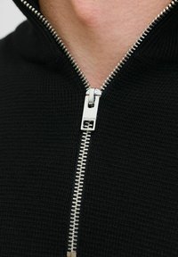 Crna zip-up dukserica s srebrnim patentnim zatvaračem i teksturiranim pletenim materijalom. Patentni zatvarač ima oznaku brenda "JACK & JONES" blizu ručice.