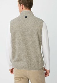 Grauer Fleece-Weste mit hohem Kragen, einer subtilen Textur und einem Logo auf der Rückseite. Kombiniert mit einem weißen Langarmshirt.