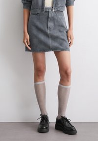 Denimskjørt med vertikale grå striper, høyt liv og knappelukking foran. Kombinert med knelengde, glitrende lysegrå sokker og svarte sko.