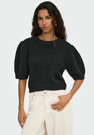 Femme aux longs cheveux foncés portant un pull noir à manches bouffantes et un pantalon blanc taille haute, debout avec une main dans la poche.