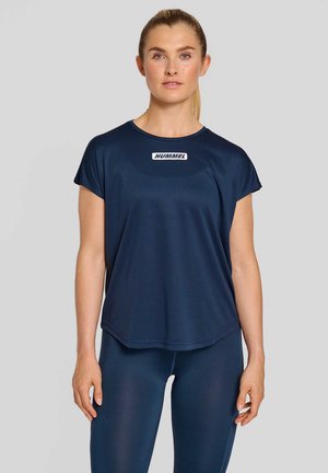 Jonge vrouw met blond paardenstaart die een marineblauw Hummel sportshirt en bijpassende leggings draagt, staand tegen een lichtgrijze achtergrond.