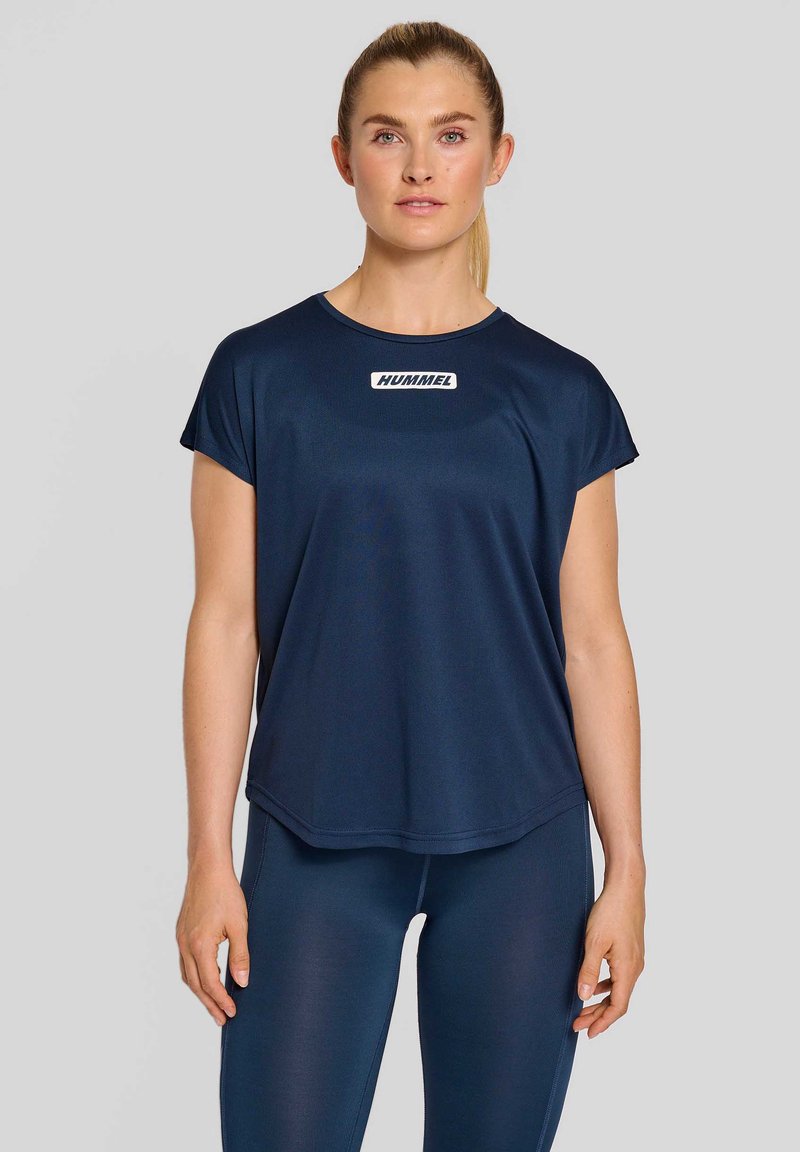 Młoda kobieta z blond kucykiem, ubrana w granatową sportową koszulkę Hummel i pasujące legginsy, stojąca na jasnoszarym tle.