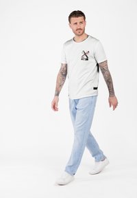 Hombre con brazos tatuados caminando, vistiendo una camiseta gráfica blanca con flores, jeans de color azul claro y zapatillas blancas, contra un fondo blanco liso.
