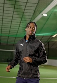 Black athletic jacket with zipper and logo, worn over a blue shirt. The setting is a sports indoor field with artificial turf.