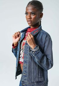 Chaqueta de mezclilla de color azul ligeramente desvaído, con botones plateados y detalles bordados en el cuello, llevada sobre una camiseta gráfica roja.