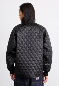 Sort quiltet jakke med diamantmønster, raglanærmer, ribbede manchetter og et logo-patch på ryggen nær taljen. Glat, satinlignende tekstur.