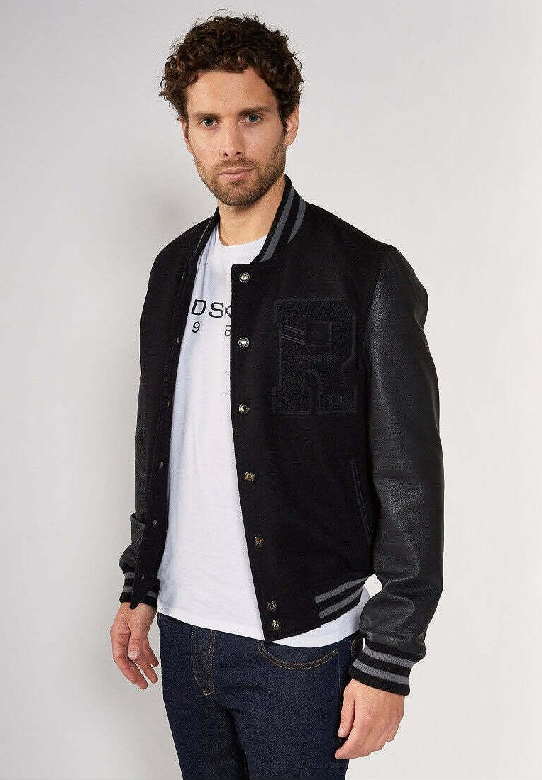 Homme aux cheveux bouclés portant une veste de lycée noire avec des manches en cuir, un t-shirt blanc imprimé et un jean bleu marine, se tenant devant un fond uni.