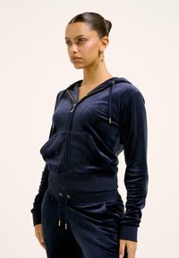 Felpa con zip in velluto blu navy con hardware dorato, tasche frontali e design aderente. Abbinata a pantaloni coordinati per un look armonioso.