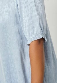Helles blaues Blusenshirt mit kurzen Puffärmel und elastischen Bündchen, das ein strukturiertes Design mit vertikalen Linien und einem glatten Stofffinish aufweist.