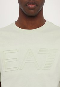 Šviesiai žalia medvilninė marškinėlių suapvalintu kaklu, turinti iškilmingą "EA7" logotipą, kuris yra šiek tiek tamsesnės spalvos. Trumpi rankoviai ir lygus paviršius.