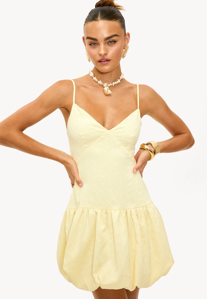 Robe jaune avec bretelles spaghetti, décolleté en V, motif floral texturé, et jupe bouffante froncée. Stylée avec des accents de bijoux.
