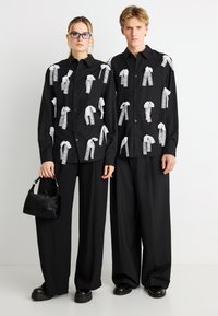 Μαύρα πουκάμισα με κουμπιά, διακοσμημένα με λευκό δαντελένιο σχέδιο, σε συνδυασμό με άνετα μαύρα παντελόνια. Το μοντέλο κρατά μια μικρή μαύρη τσάντα. Ομοιόμορφη σχεδίαση και υφή.