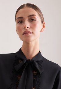 Blusa nera con collo a fiocco, dettagli in pizzo raffinati sulla parte anteriore e tessuto liscio e strutturato.