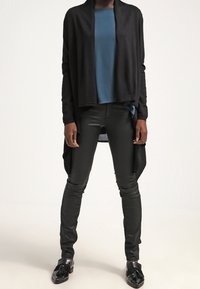 Černý cardigan s otevřeným předním dílem přes modrý top, doplněný úzkými černými koženými kalhotami a černými botami s mašlí. Uklizený, štíhlý silueta.