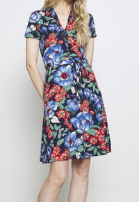 Donna che indossa un vestito blu navy al ginocchio con grandi stampe floreali blu e rosse, un collo a V con bottoni, maniche corte e una cintura annodata in vita.