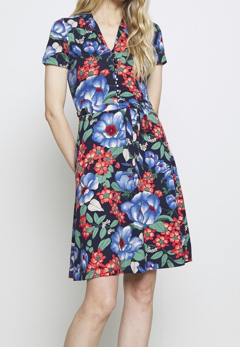 Donna che indossa un vestito blu navy al ginocchio con grandi stampe floreali blu e rosse, un collo a V con bottoni, maniche corte e una cintura annodata in vita.
