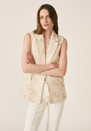 Mujer con cabello largo y castaño, vistiendo un blazer sin mangas beige con bordados florales y pantalones blancos, con los brazos cruzados frente a un fondo liso.